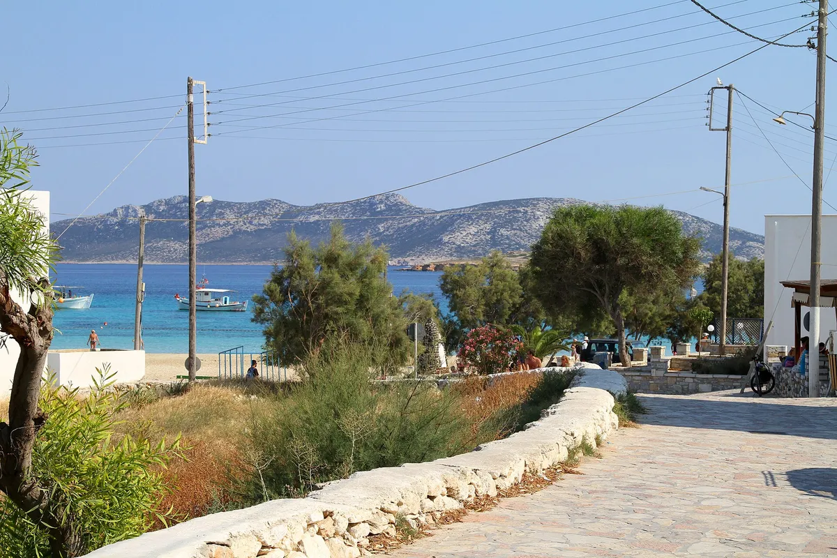 Village path by a low stone wall leading toward a turquoise bay with fishing boats on Koufonisia, Greece
