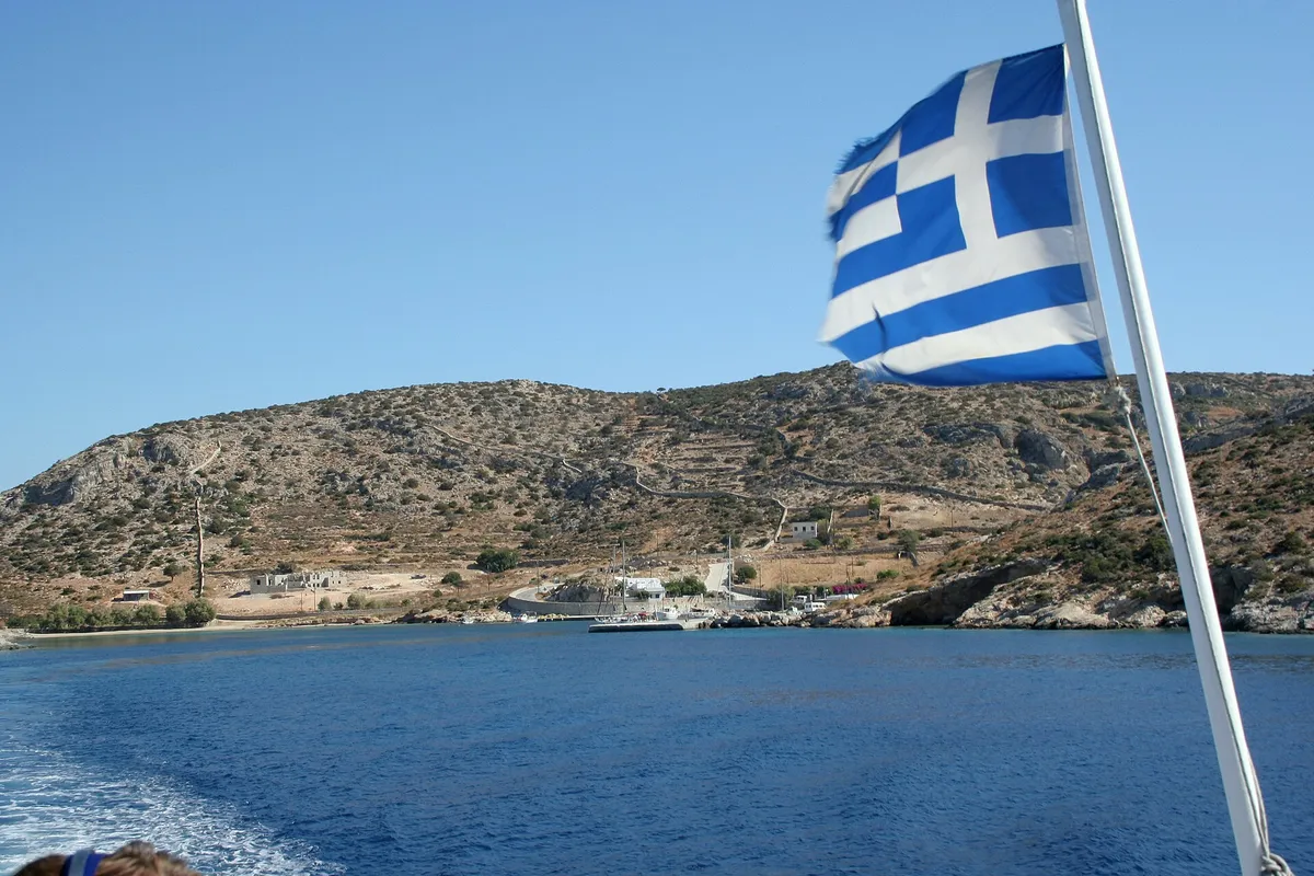 Greek flag on a boat approaching a Small Cyclades harbour with hills and moored boats, on a boat trip from Naxos